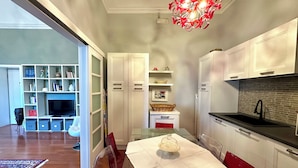 Una cocina moderna con armarios blancos, encimera negra y una silla roja. Hay una mesa de comedor con cubierta de vidrio y mantel blanco. Un candelabro rojo cuelga del techo.