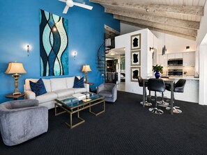 A modern living room with a blue accent wall, a glass coffee table, and a dining area with bar stools. - Sunset Serenade: Inspiring Views, Modern Desert Sanctuary...managed by Greenday! (Rancho Mirage)