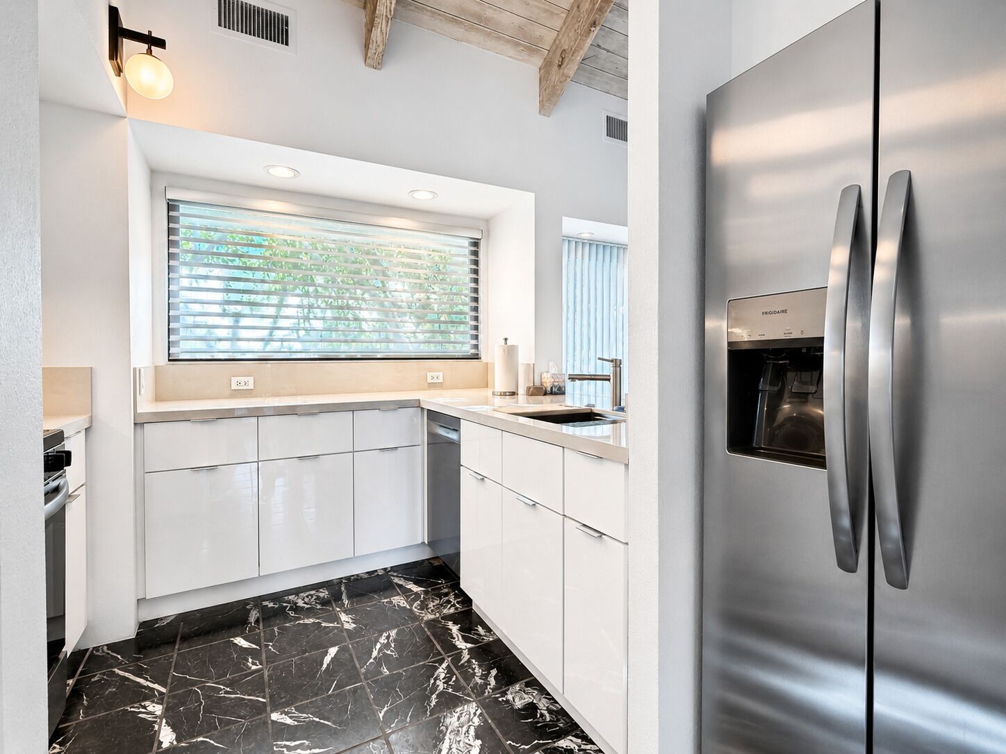 A modern kitchen with a built-in refrigerator, white cabinetry, a window with blinds, and a marble floor.