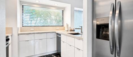 A modern kitchen with a built-in refrigerator, white cabinetry, a window with blinds, and a marble floor.