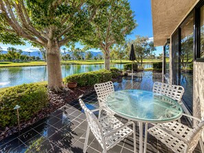 A glass-top table with white wicker chairs on a patio overlooking a lake, with a golf course and trees in the background. - Sunset Serenade: Inspiring Views, Modern Desert Sanctuary...managed by Greenday! (Rancho Mirage)