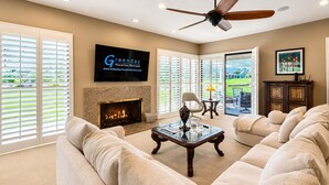 A spacious living room with a fireplace, a ceiling fan, and a large TV mounted on the wall. - Harvard Hideaway: sun-splashed, westerly views in The Springs Country Club. Managed by Greenday! (Rancho Mirage)