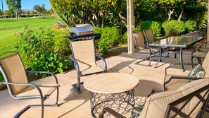 Eine Terrasse im Freien mit einem runden Tisch, Stühlen, einem Grill und Blick auf einen Golfplatz.
