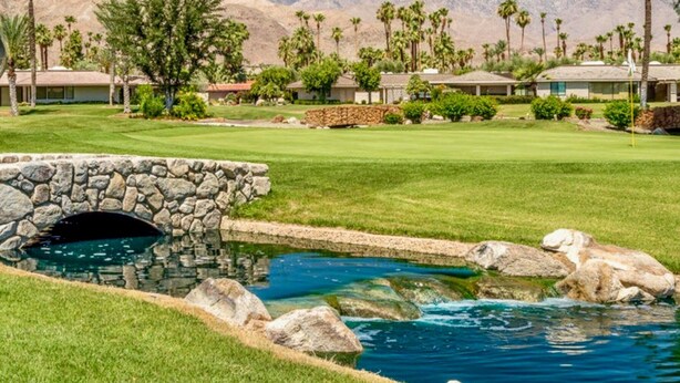 A golf course with a stone bridge over a pond, surrounded by palm trees and buildings with mountain views in the background. - Harvard Hideaway: sun-splashed, westerly views in The Springs Country Club. Managed by Greenday! (Rancho Mirage)