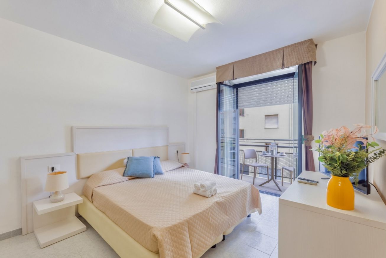Habitació d'hotel amb llit, taulella de nit, làmpada i vista al balcó.