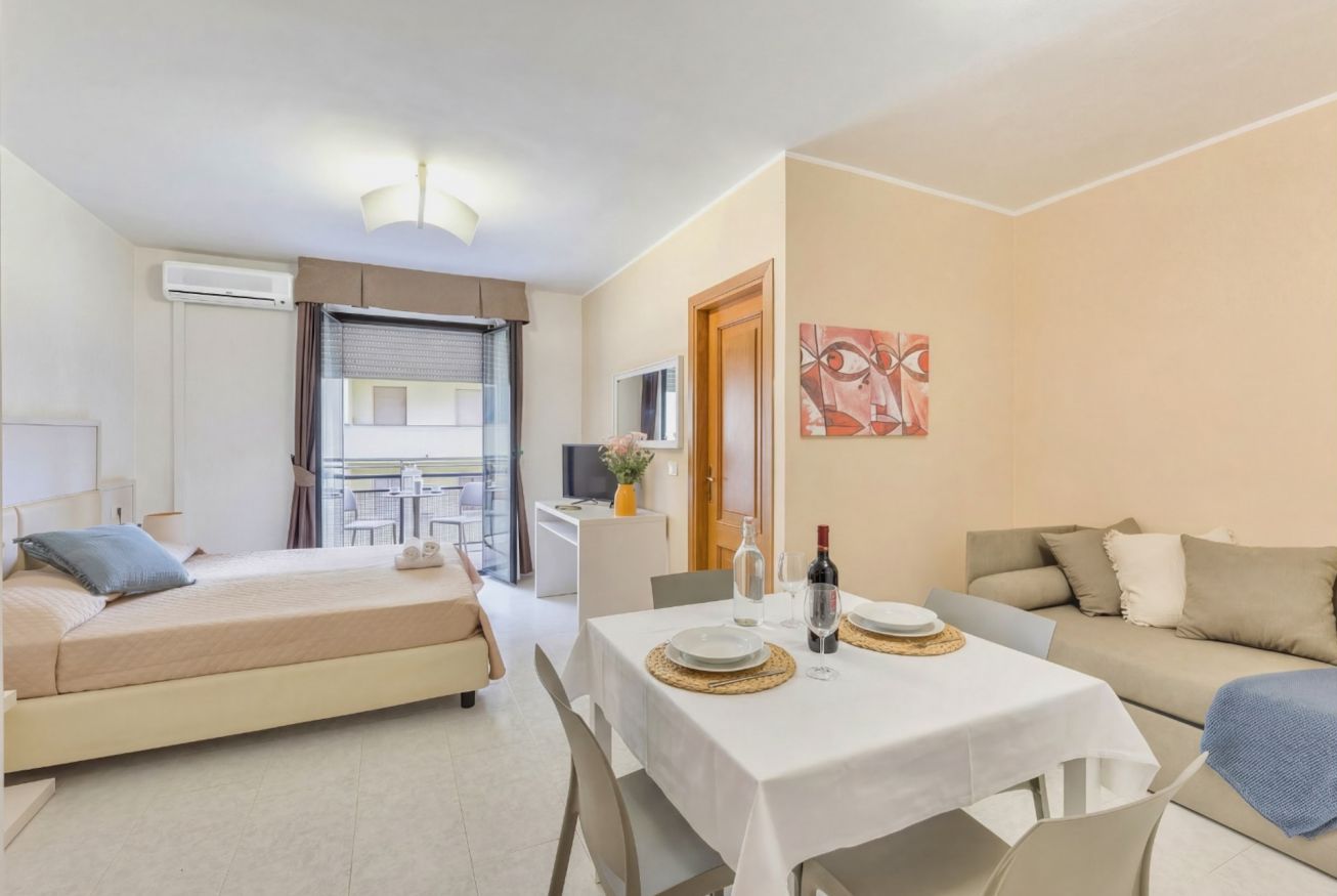 Una camera d'albergo moderna con un letto, un tavolo da pranzo per due persone, un divano e un balcone con tavolo e sedie.