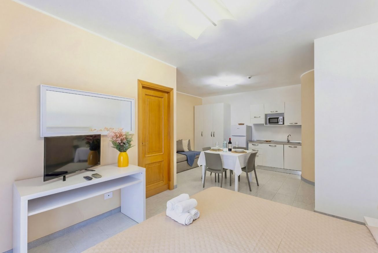 Quarto de hotel moderno com televisão, porta de madeira, área de refeições com mesa e cadeiras, e cozinha compacta.