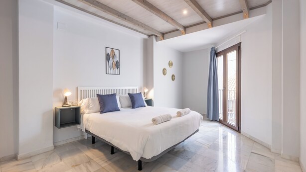 A bedroom with a large bed, a nightstand, a wall-mounted light, and a window with curtains. - Apartamentos Realejo Granada Centro 3000 (Granada)