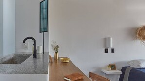 Una cocina moderna con encimera de granito, grifo negro y un florero con flores. Se observa una mesa de comedor de madera con un libro y un tazón. Al fondo, se ve una cama con manta azul y almohada blanca.