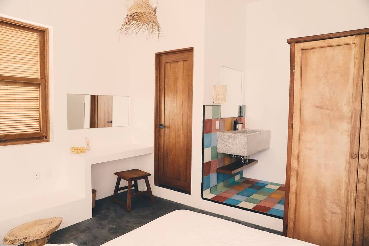 A bedroom with a bed, a wooden door, a small stool, a mirror, and a colorful tiled area.