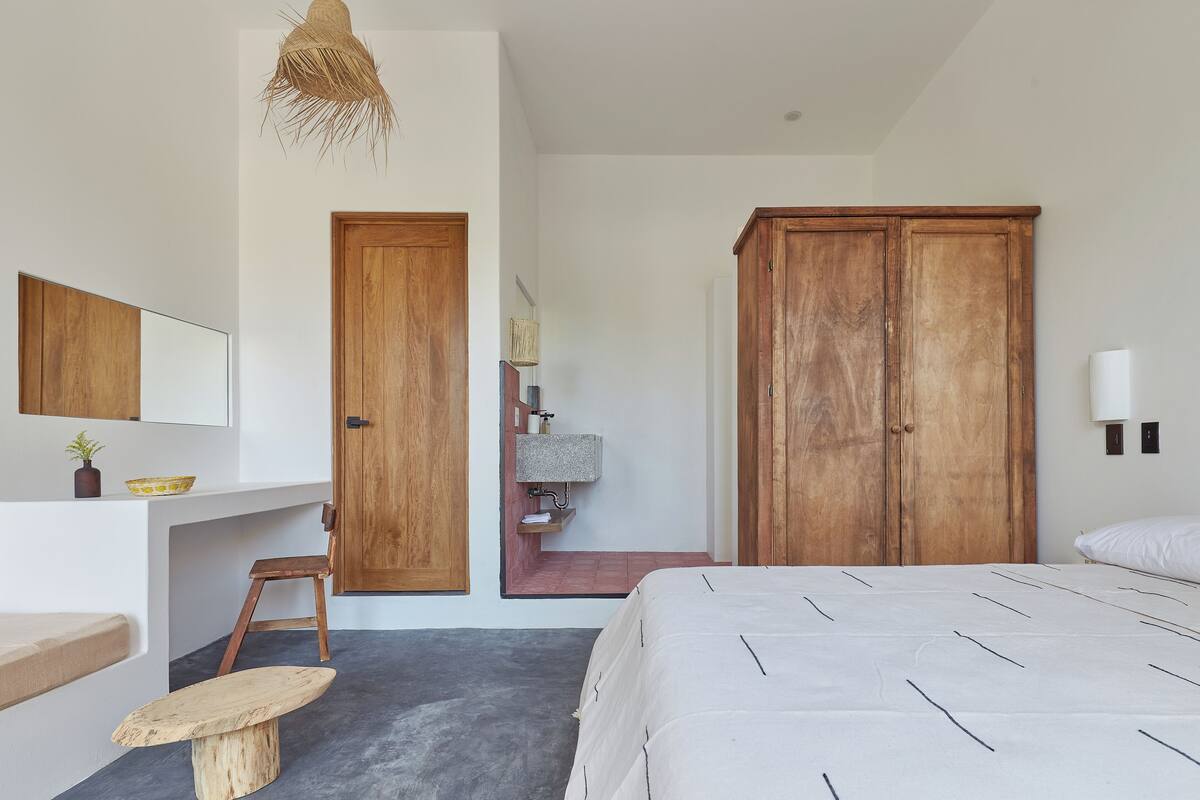 A bedroom with a bed, wooden wardrobe, a desk with a chair, a small stool, a hanging light fixture, and a wall-mounted shelf with a mirror and a plant.