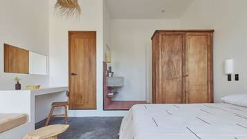 Un dormitorio con cama, armario de madera, escritorio con silla, taburete pequeño, lámpara colgante y estante con espejo y planta.