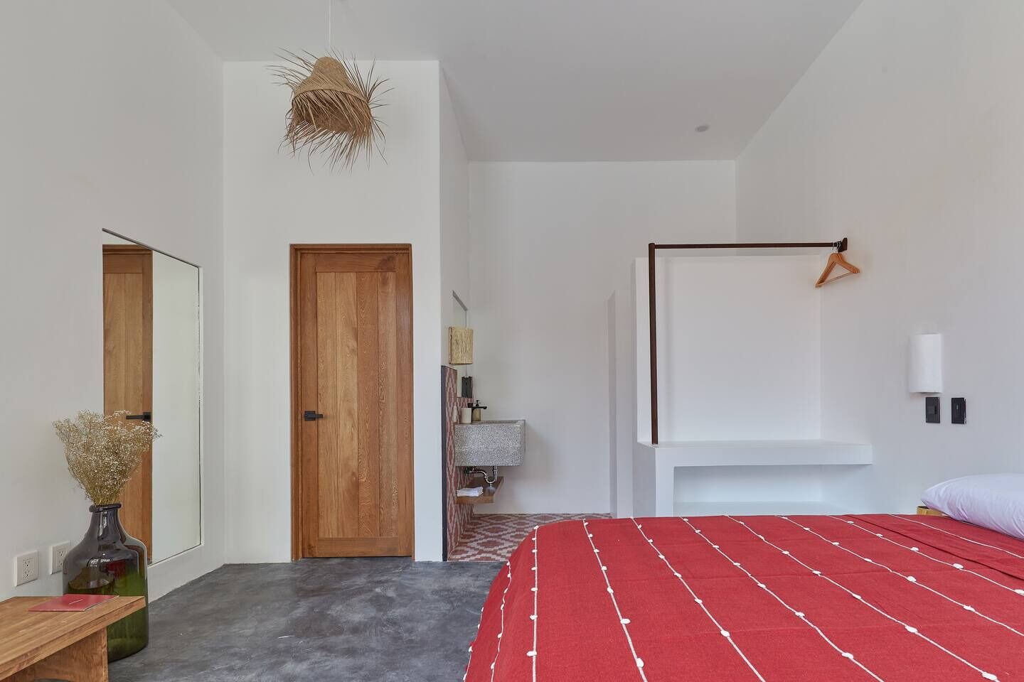 Une chambre moderne avec une porte en bois, une couvre-lit rouge, un luminaire suspendu et une petite table de chevet sur laquelle est posé un vase.