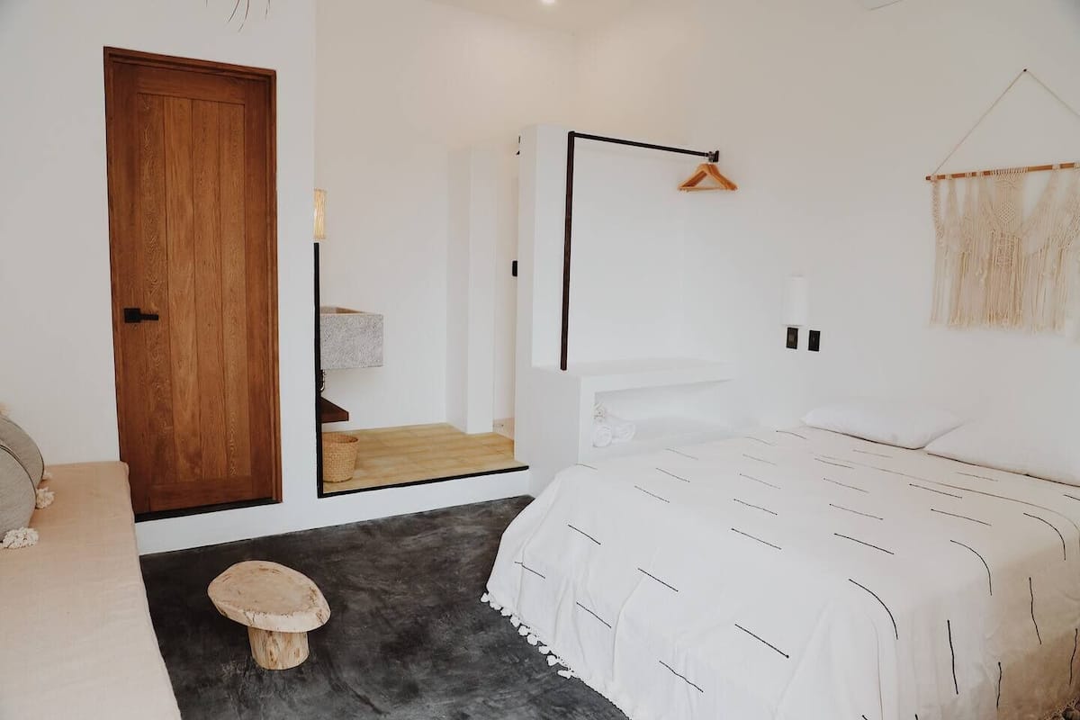 A minimalist bedroom with a wooden door, a bed with a white bedspread, a small stool, and a hanging macrame piece.