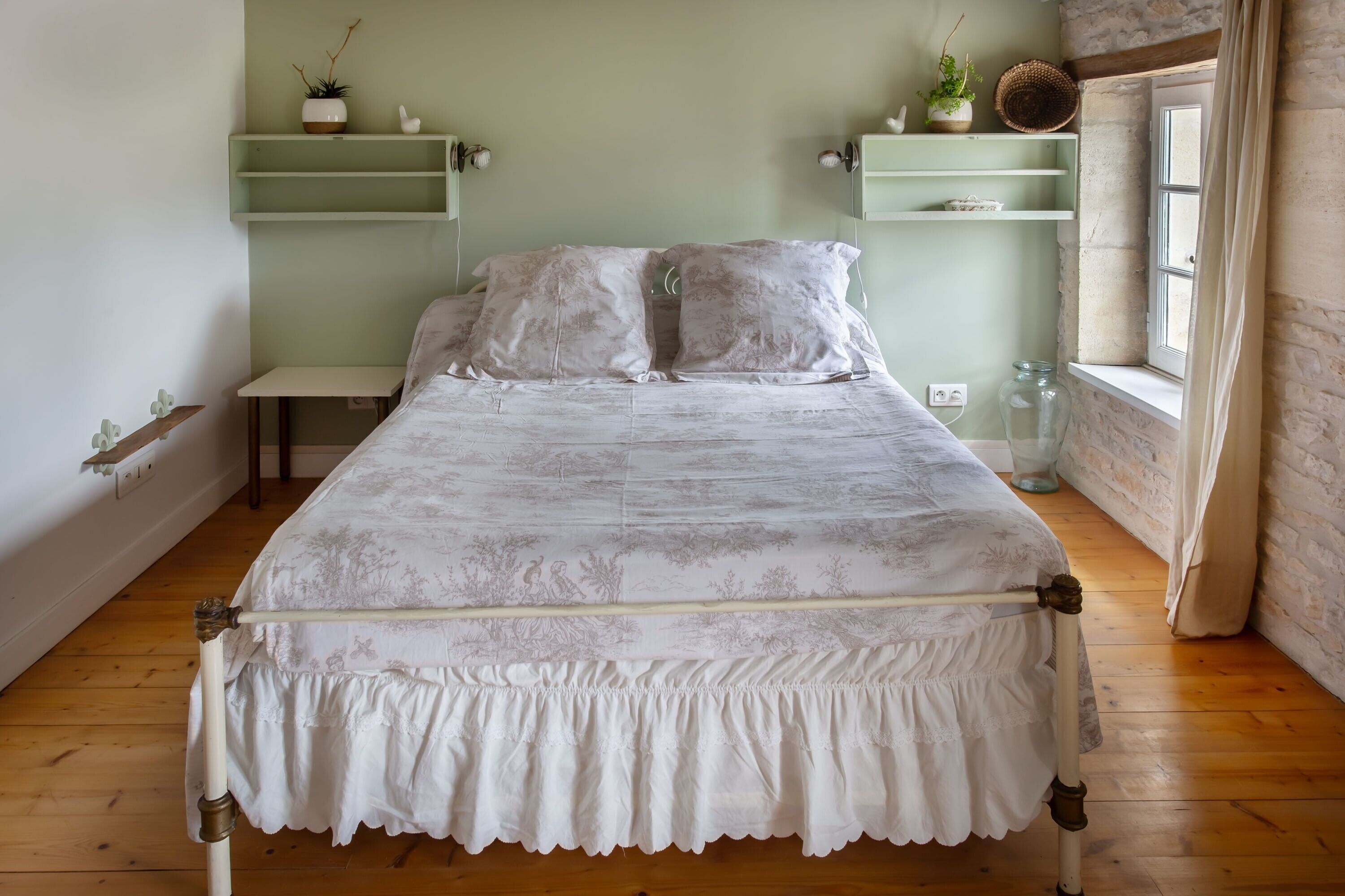 Ein Bett mit weißer, floraler Bettdecke, Holzboden und einer grünen Regalwand mit Pflanzen.