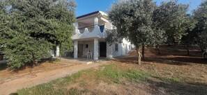 Une maison à deux étages avec un balcon, entourée d’oliviers et bordée d’un chemin de terre.