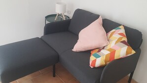 A dark grey sofa with a matching ottoman, a white lamp on a small side table, and a colorful geometric pillow. - Vacation home 200 mt from the sea in sardinia (Tertenia)