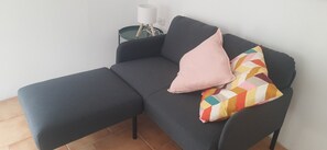 Un canapé gris foncé accompagné d’un pouf assorti, une lampe blanche posée sur une petite table d’appoint et un coussin aux motifs géométriques et aux couleurs vives.