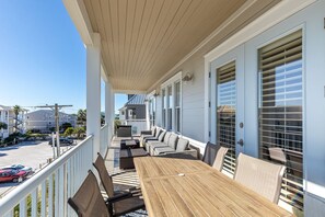 Un balcon couvert avec une table et des chaises en bois, donnant sur la rue, et un immeuble en arrière-plan.