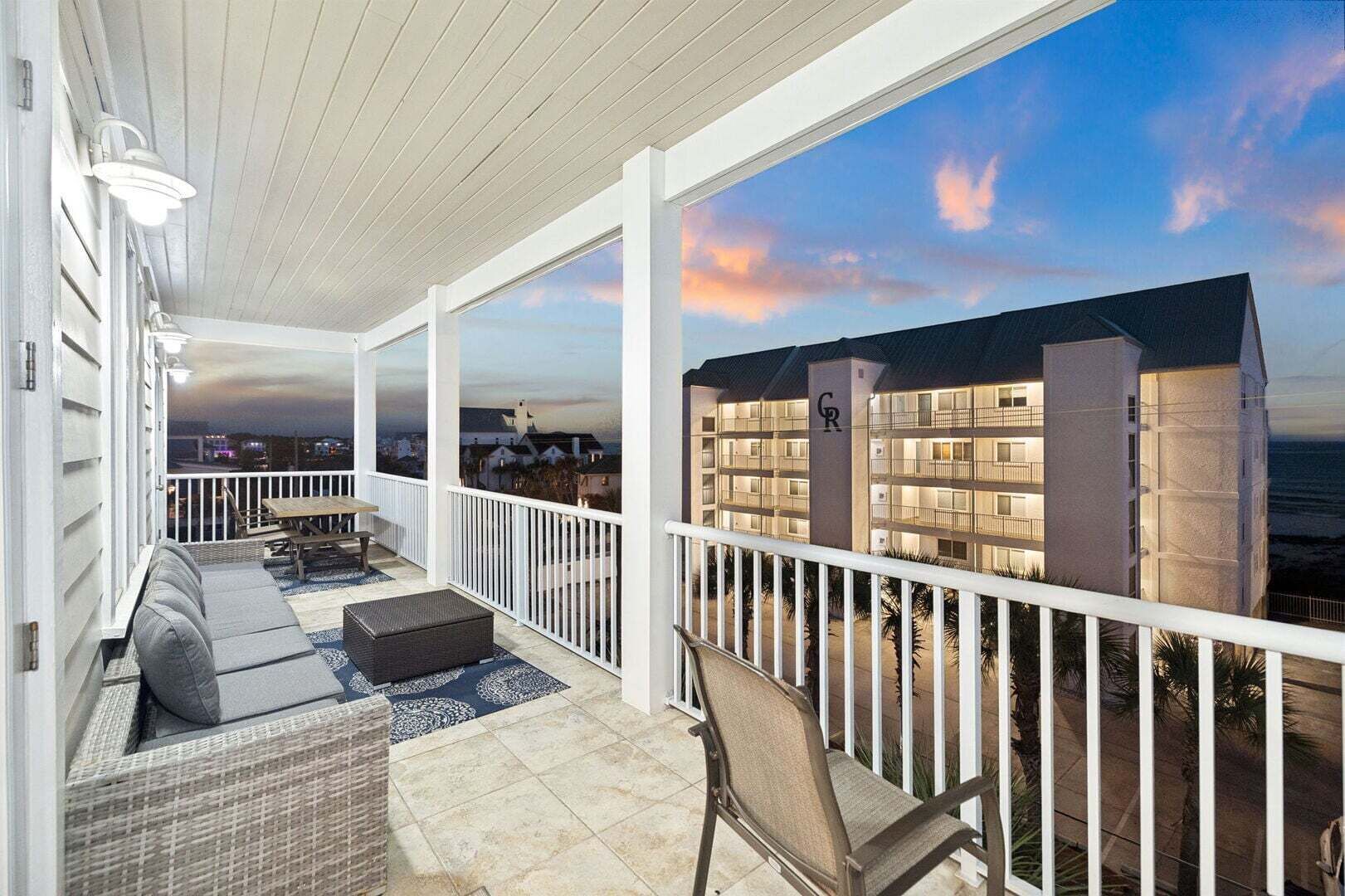 A covered balcony with wicker furniture, a view of a building with balconies, and a sunset sky.