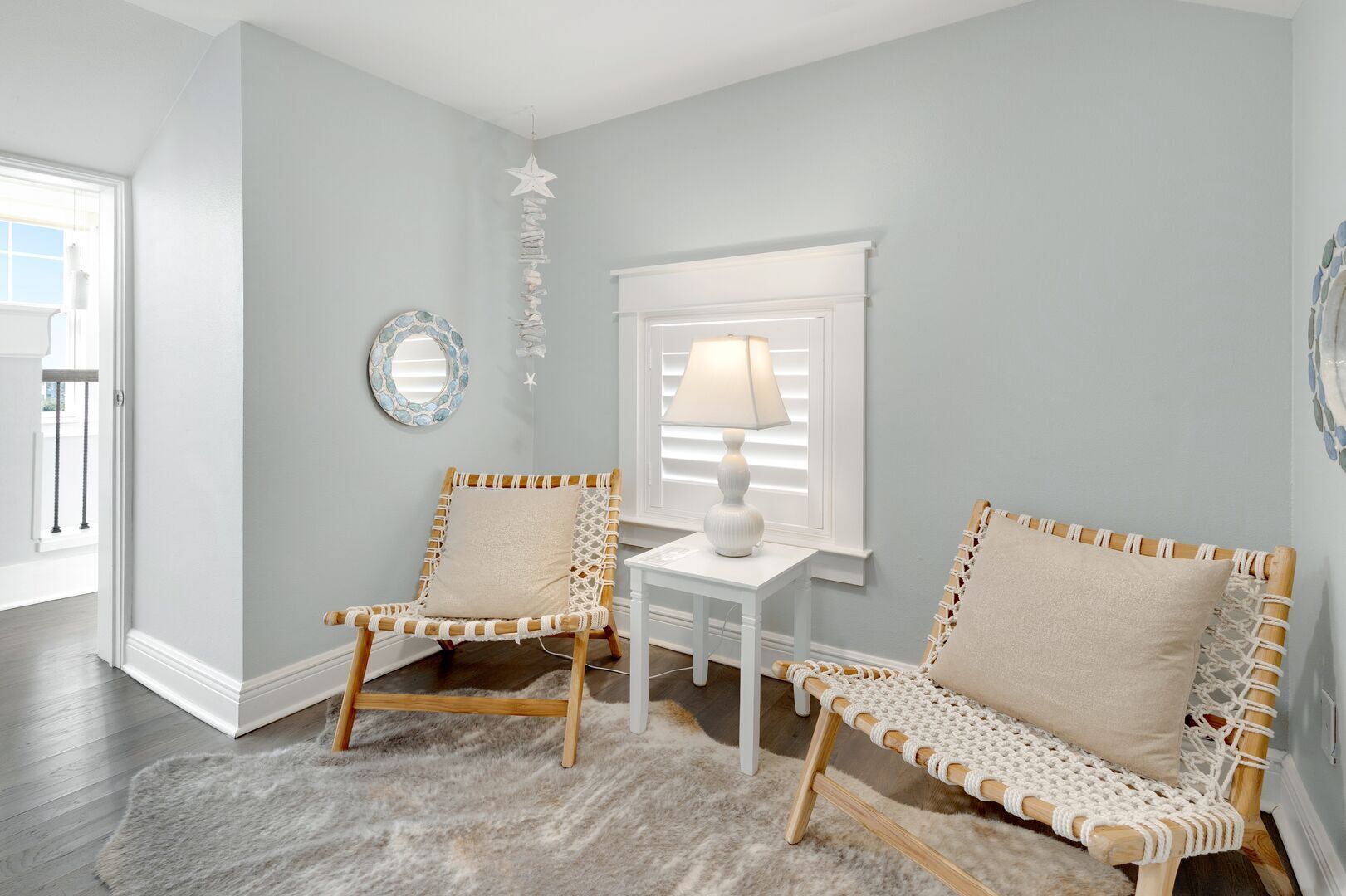 A room with two wicker chairs, a small white table with a lamp, and a mirror on the wall.