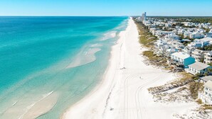 En kuststad med en lång sandstrand, kristallklart turkost vatten och hus längs stranden med blå tak.