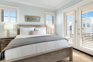 Une chambre avec un lit en bois, du linge de lit blanc, un tableau accroché au mur et une vue à travers les stores de la fenêtre.