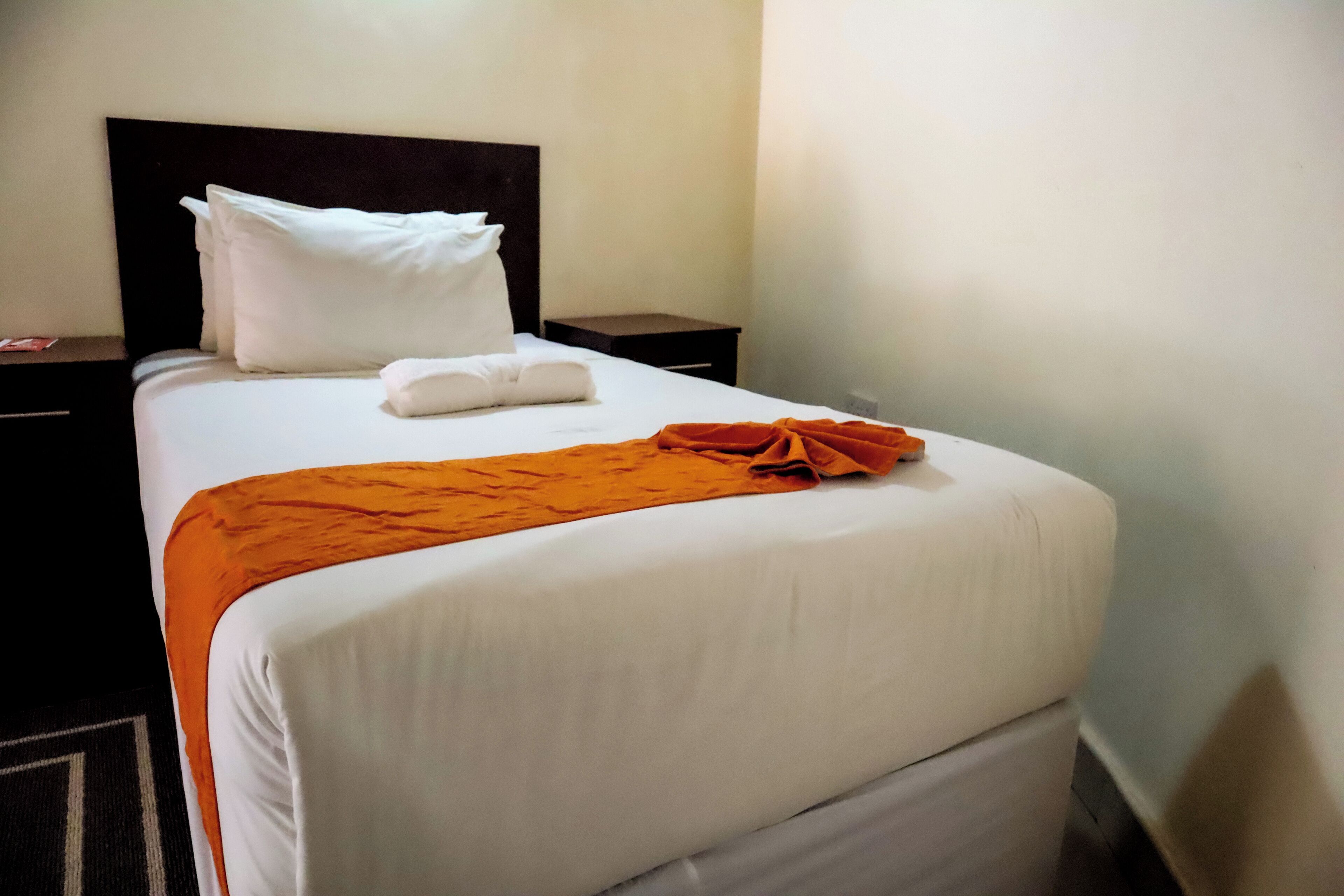 A neatly made bed with white linens, a dark headboard, and an orange throw blanket.