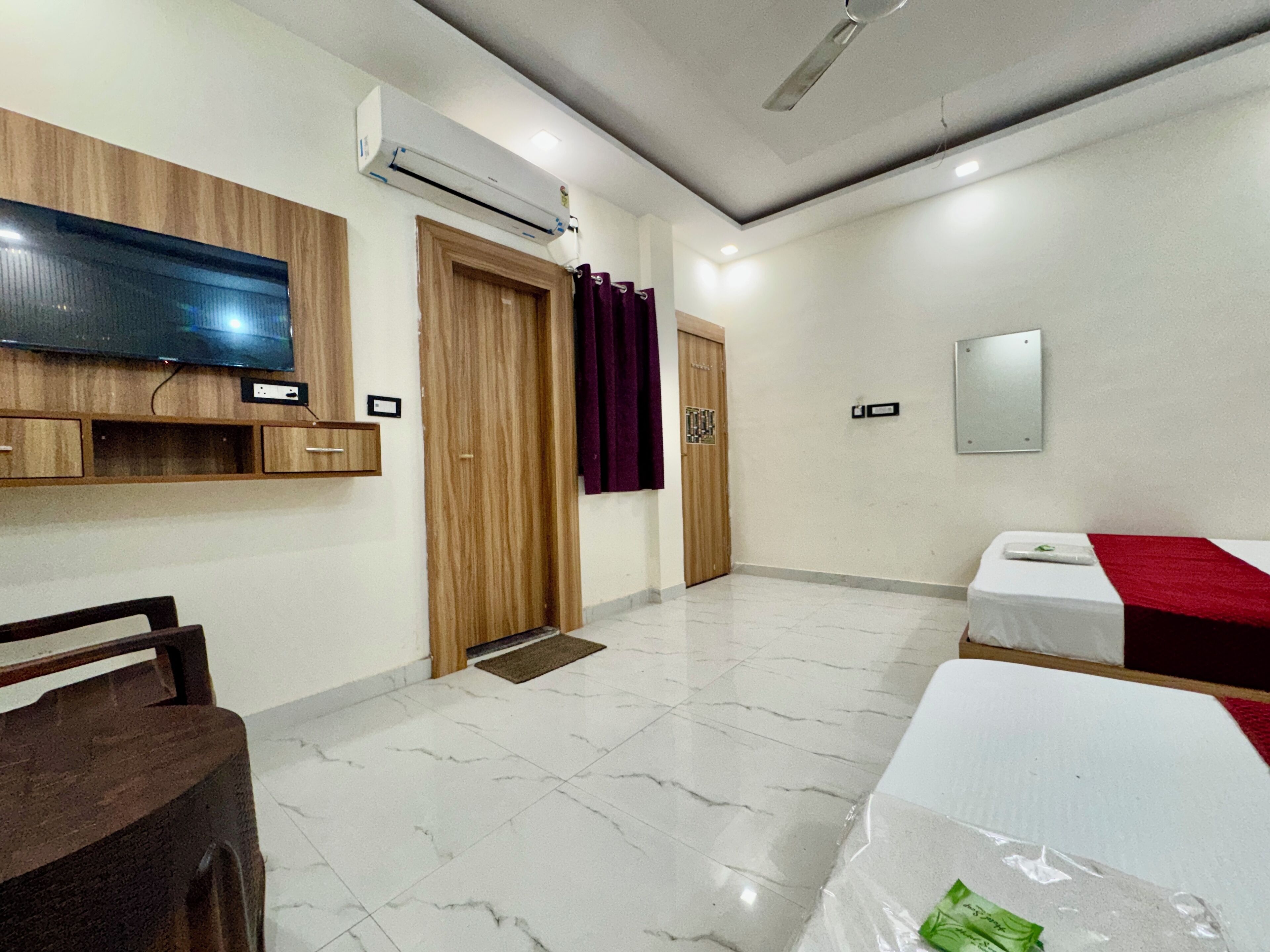 A hotel room with a flat-screen TV, wooden wardrobe, two beds, a mirror, and a ceiling fan.