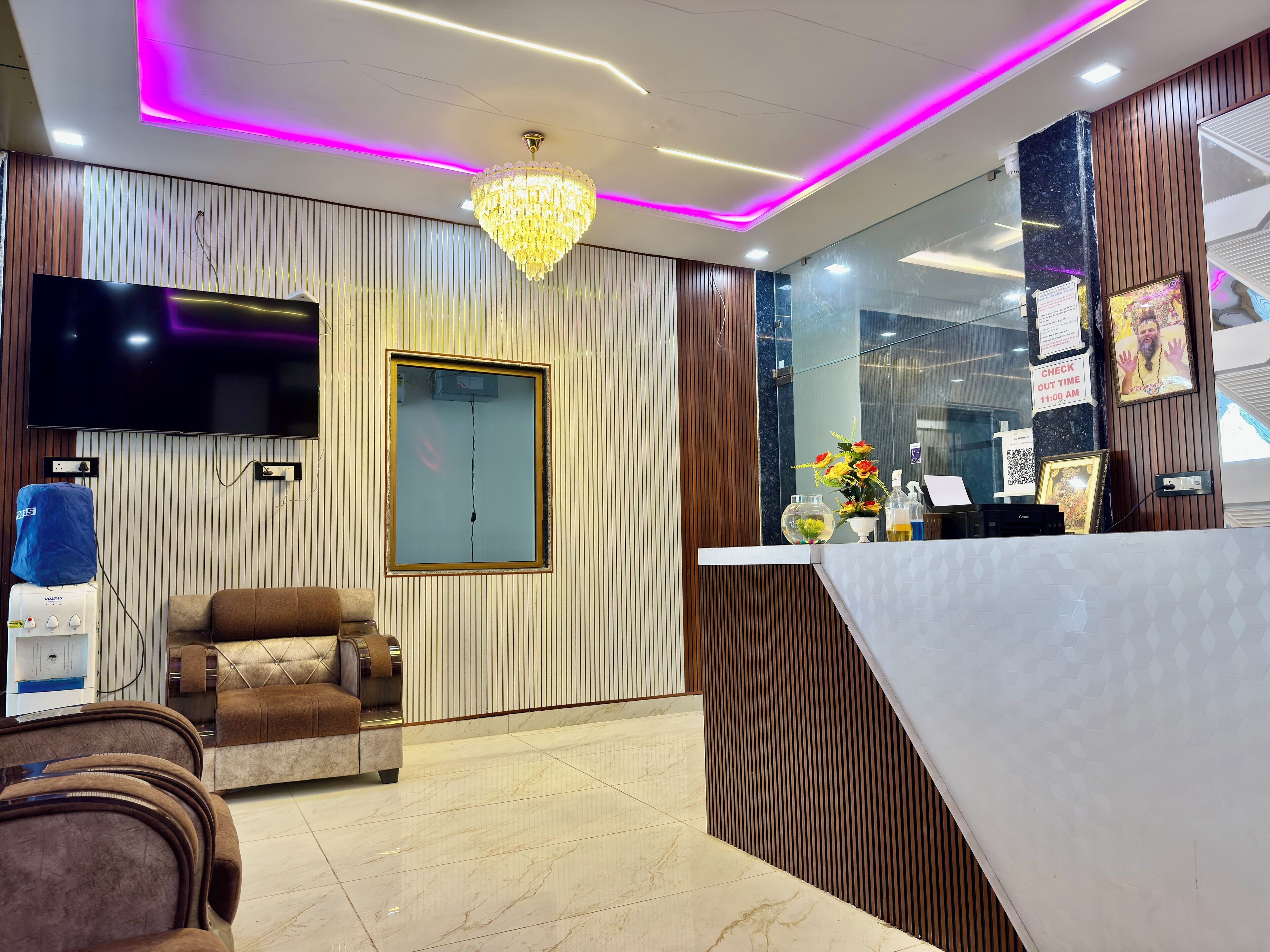 A reception area with a modern chandelier, a flat-screen TV, a wooden reception desk, and a seating area with a brown leather sofa.
