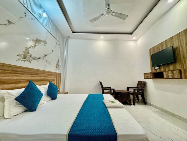 A hotel room with a bed, a ceiling fan, a TV mounted on the wall, and a small table with chairs. - Hotel Krishna Kripa Palace (Vrindavan)