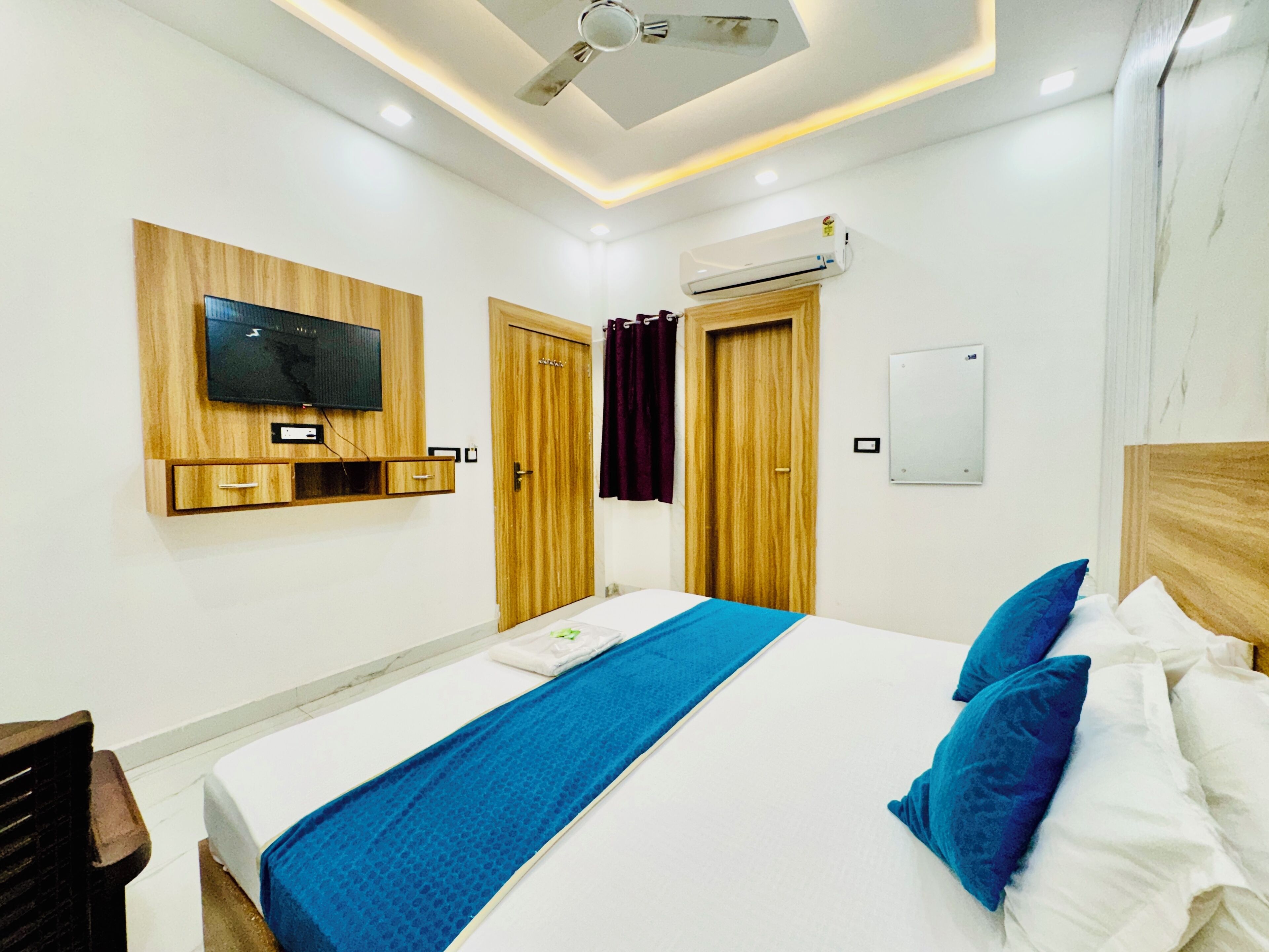 A hotel room with a bed, a TV mounted on the wall, an air conditioning unit, and wooden doors.