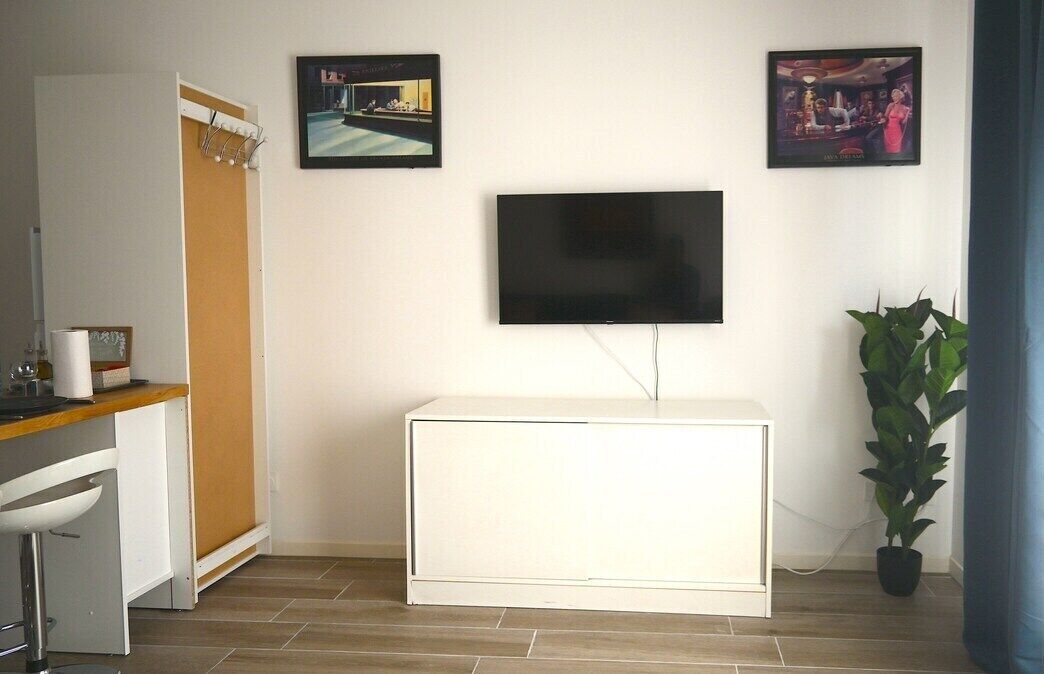 Una sala moderna con televisor de pantalla plana montado en la pared, mueble blanco para entretenimiento, zona de comedor con taburete de bar y una planta en maceta.