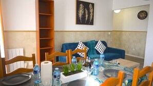 Uma área de refeições com uma mesa posta para dois, um sofá azul, uma estante de madeira e um relógio de parede.