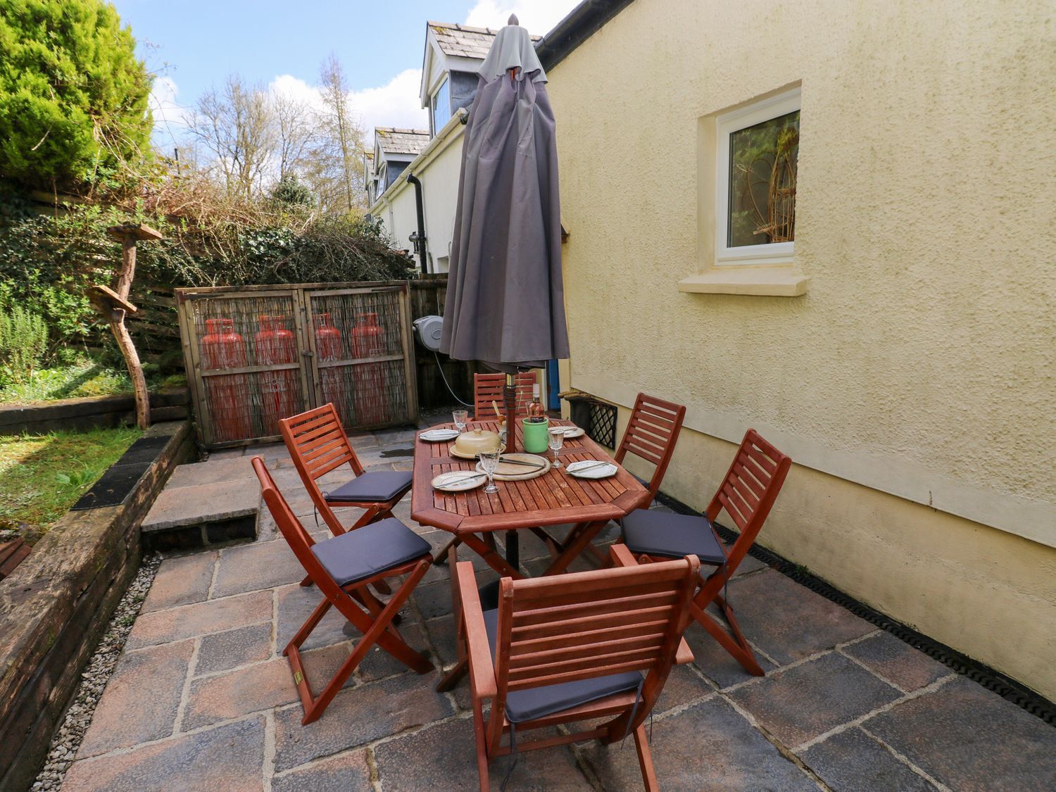 A patio with a wooden table and chairs, a grey umbrella, and a small window.
