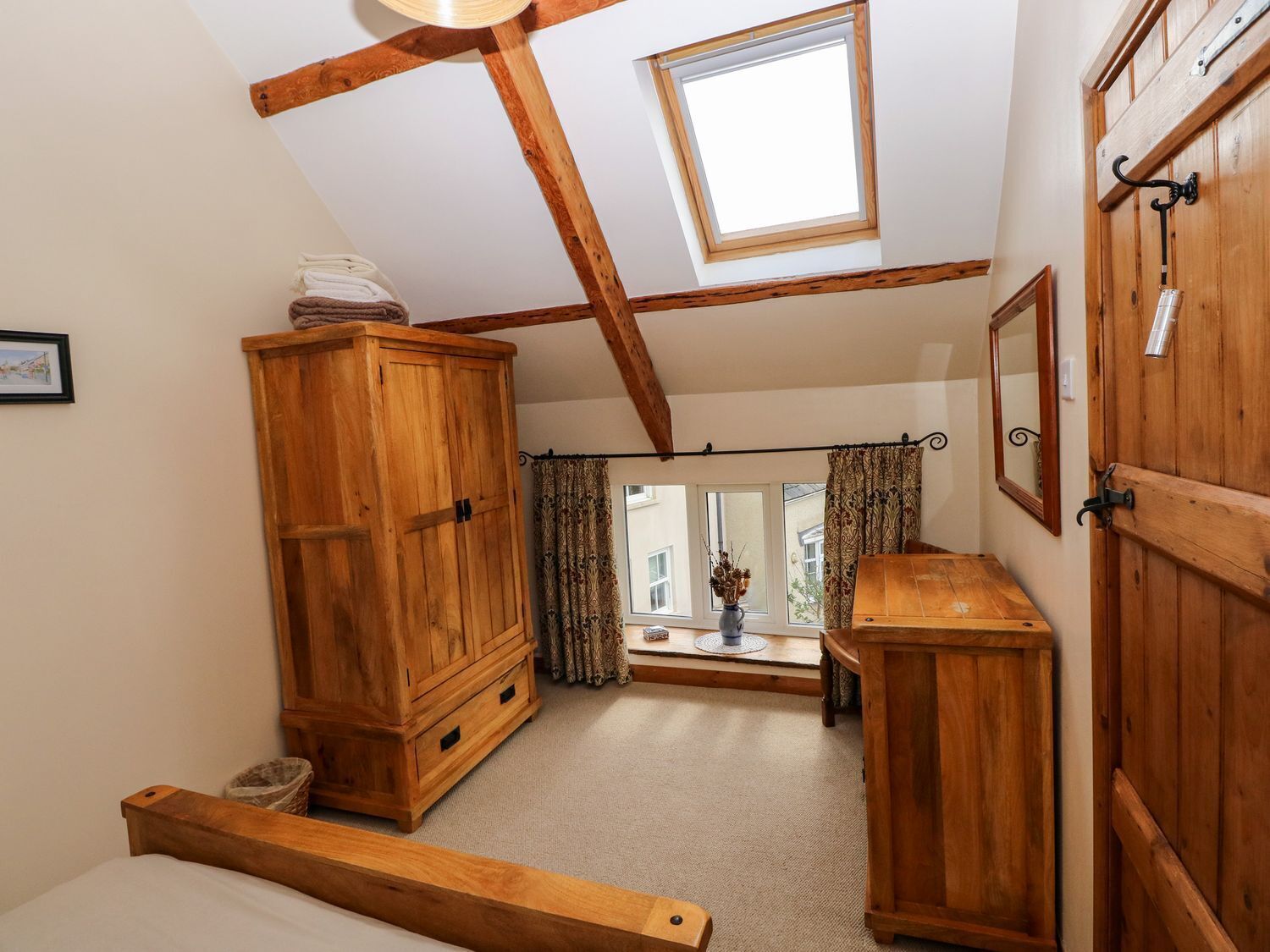 Une pièce avec un plafond en pente, des poutres en bois, une lucarne, une armoire en bois, une petite table avec un vase et une fenêtre avec des rideaux.