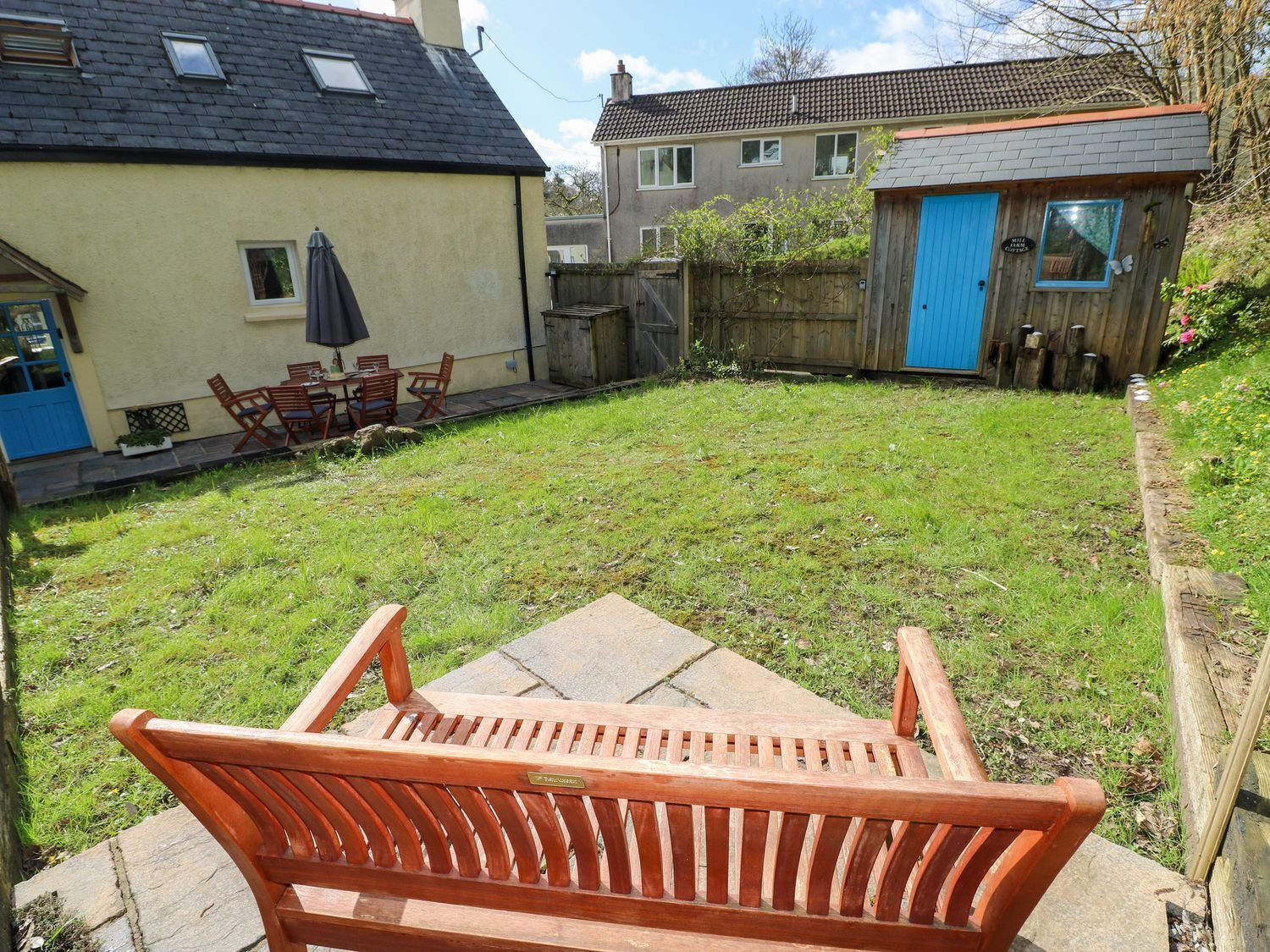 Un banc en bois dans un jardin, avec une maison, un abri de jardin et une table de patio entourée de chaises.
