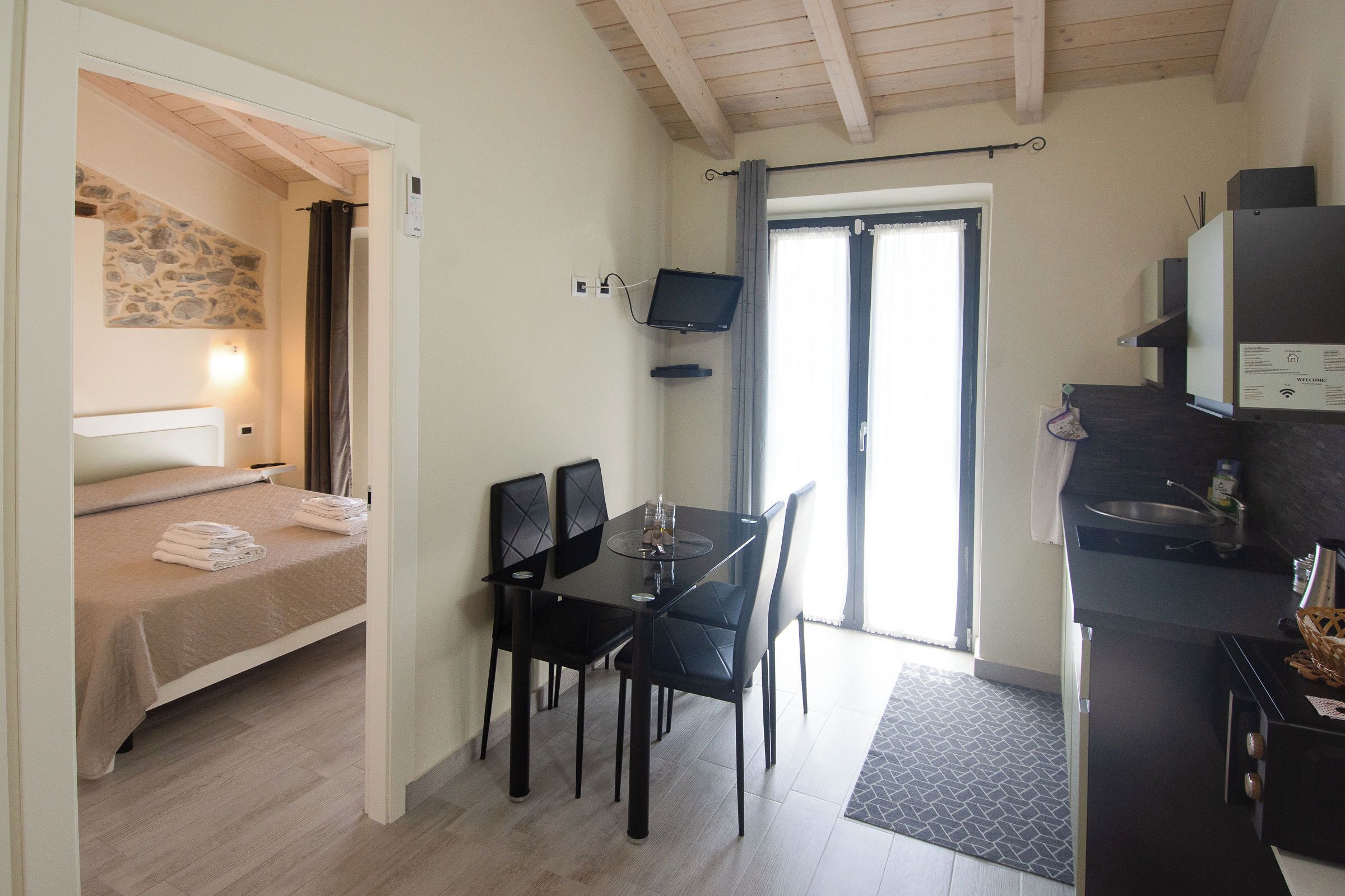 Una habitación compacta con cama, mesa de comedor con sillas, pequeña área de cocina y televisor montado en la pared.