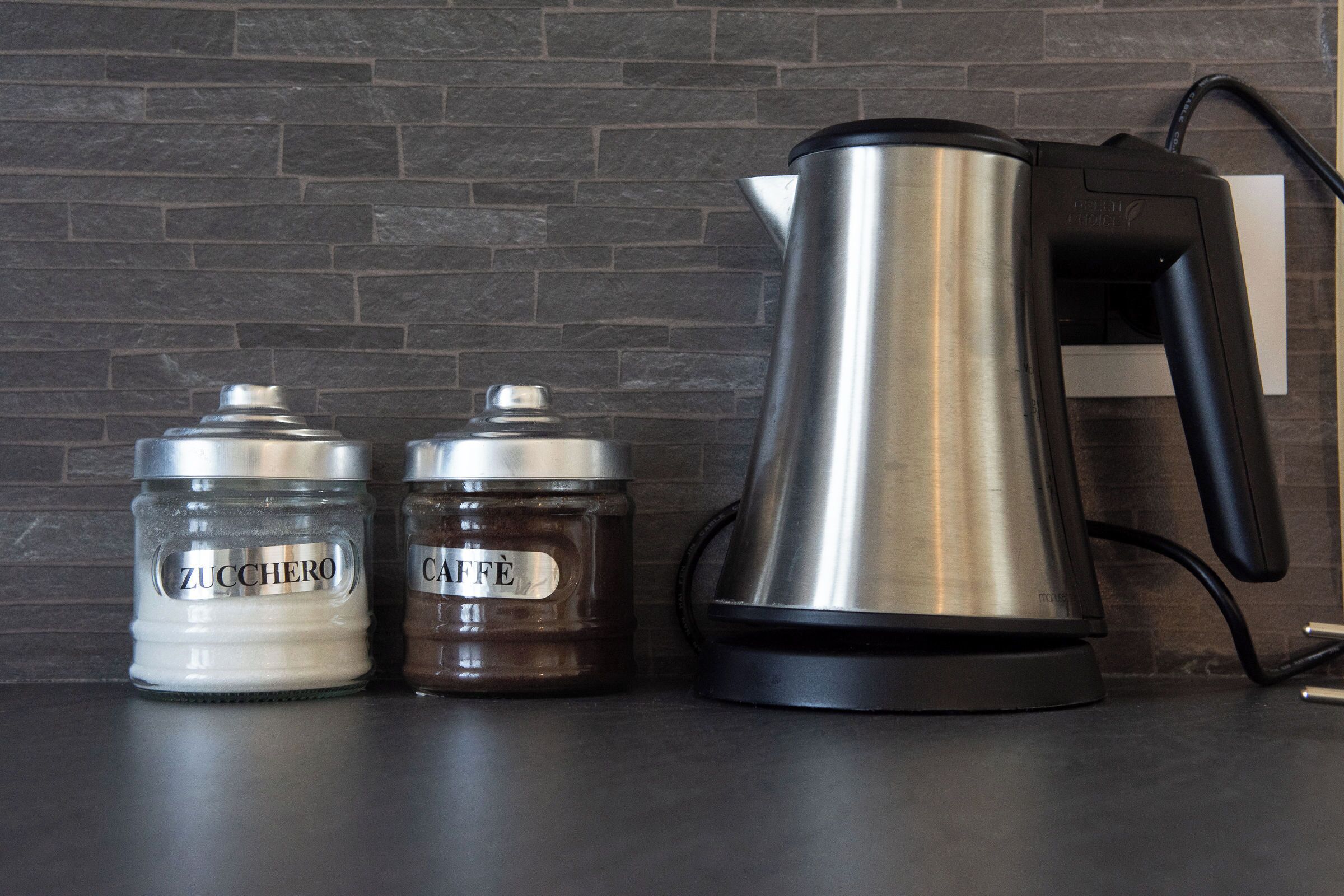 Un comptoir de cuisine avec une machine à café, deux bocaux étiquetés « ZUCCHERO » et « CAFFÈ », et un mur en briques en arrière-plan.