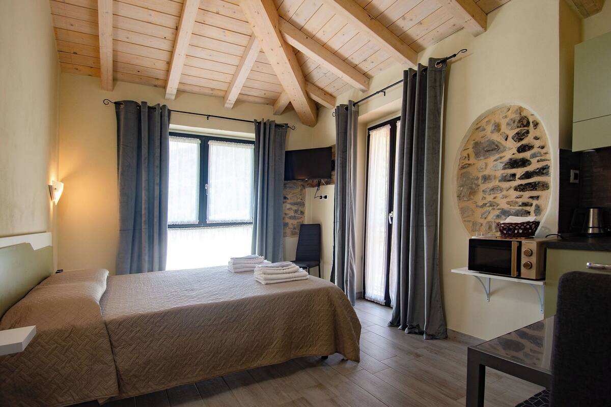 Un dormitorio con cama, cortinas, una pared de piedra, un microondas y una mesita.