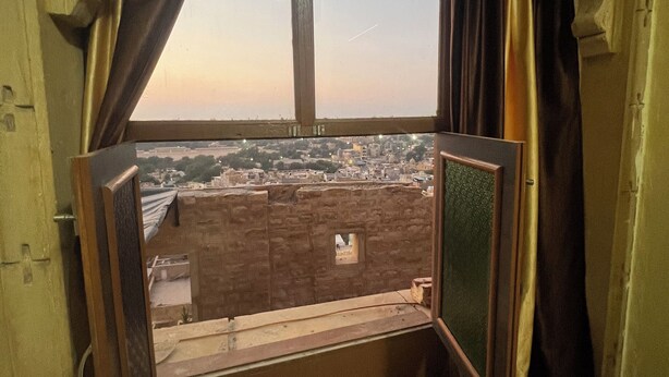 A room with a window overlooking a cityscape, featuring a stone wall and a small balcony. - La Purezza stay and restaurant  (Jaisalmer)