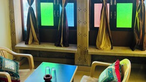 A room with two windows, each with curtains tied back, revealing a green screen. There is a table with a cushion and a chair with a patterned cushion. - La Purezza stay and restaurant  (Jaisalmer)