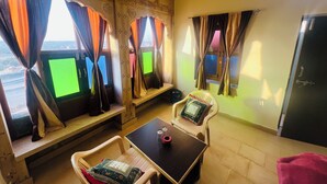 A room with a view of a bridge, a table with a red object, and two chairs with patterned cushions. - La Purezza stay and restaurant  (Jaisalmer)