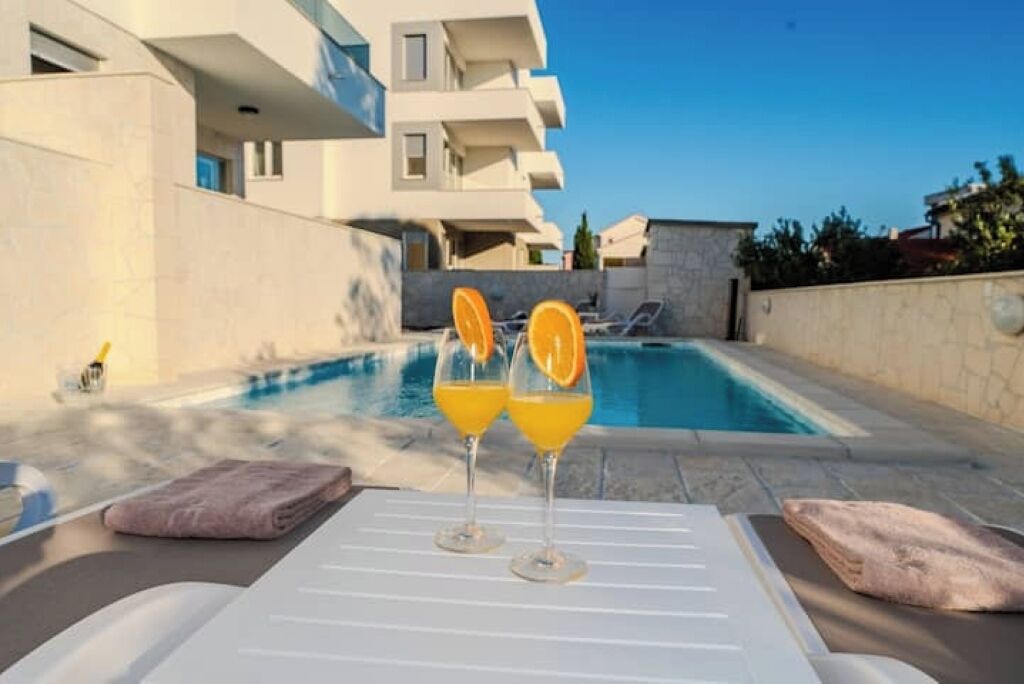 A poolside area with lounge chairs and a table, two glasses of orange juice, and a view of a modern building with balconies.