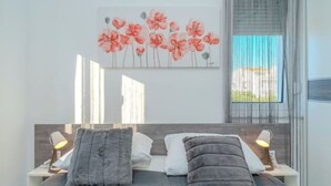 Ein modernes Schlafzimmer mit einem Bett, zwei Nachttischen mit Lampen, einem großen Fenster mit durchsichtigen Vorhängen und einem Blumenbild an der Wand.