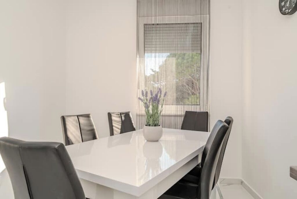 Uma sala de jantar moderna com uma mesa branca e cadeiras pretas, uma planta em vaso e uma janela com cortinas leves.