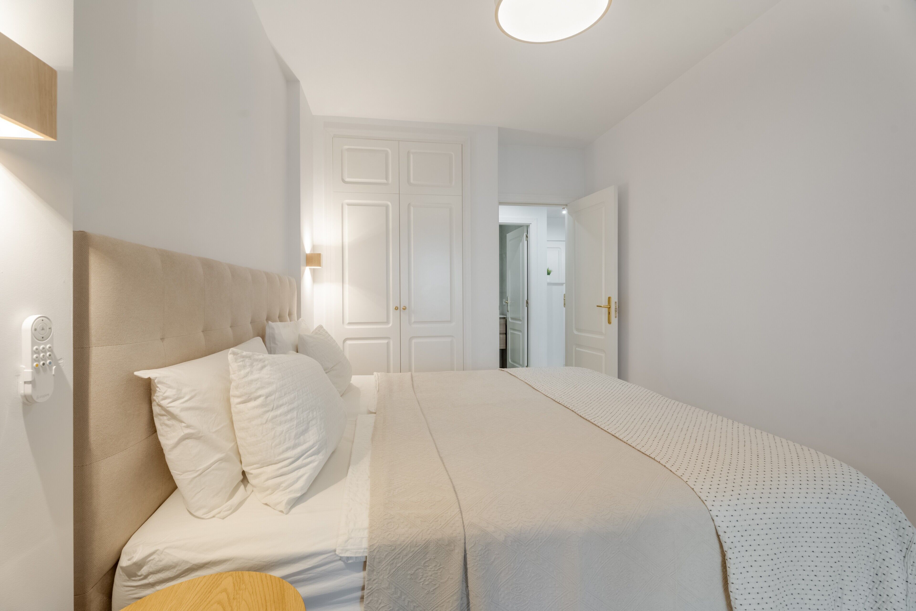 Un dormitorio con cama, mesita de noche, luz empotrada en la pared y una puerta que da a otra habitación.