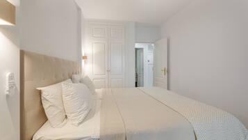 Un dormitorio con cama, mesita de noche, luz empotrada en la pared y una puerta que da a otra habitación.