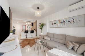 Un salon moderne avec un canapé, une table basse et un téléviseur. On aperçoit à l’arrière-plan un coin repas agrémenté d’une plante et d’un tableau accroché au mur.