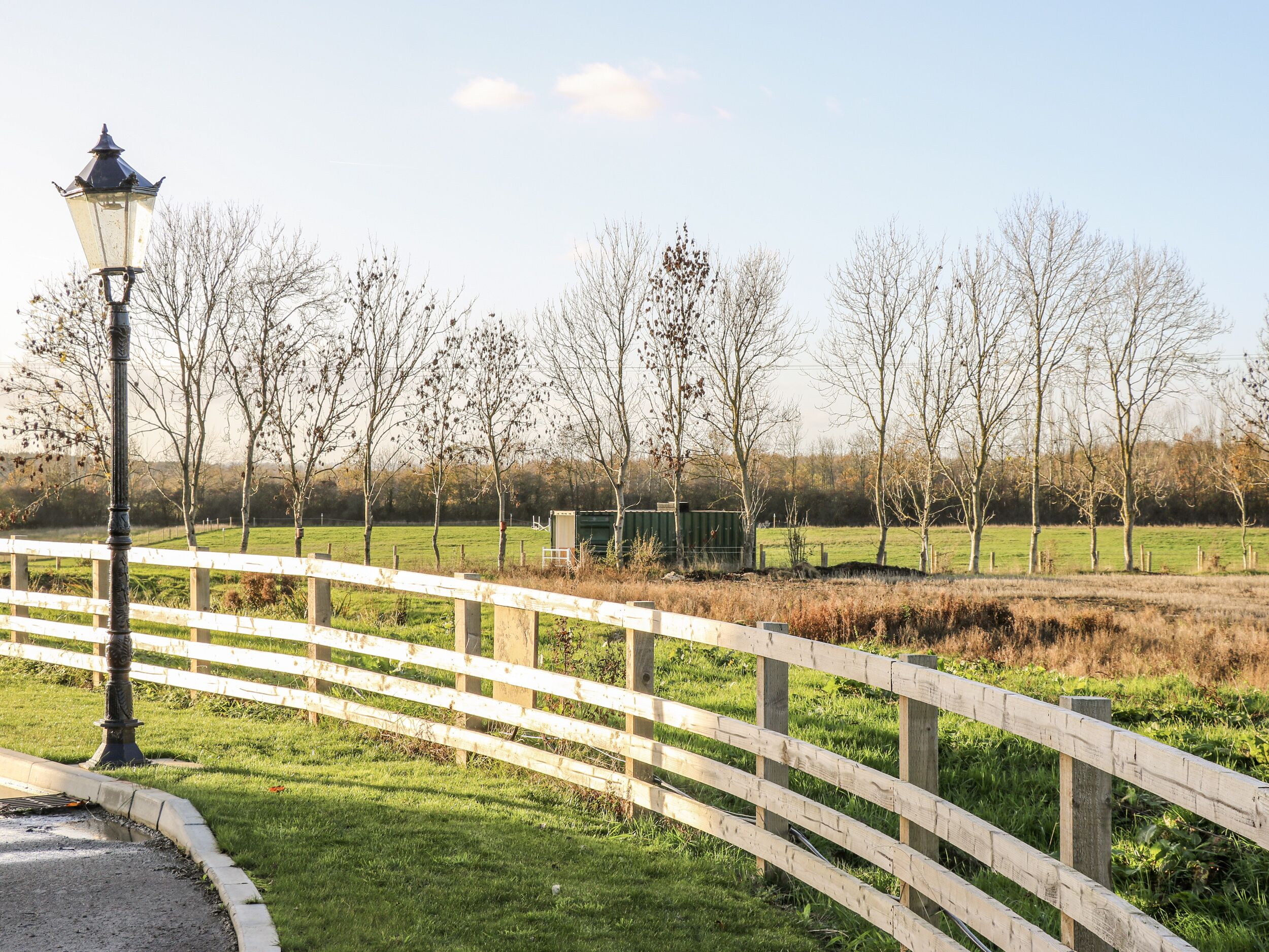 Een houten hek, een lantaarnpaal en kale bomen in een grasveld.